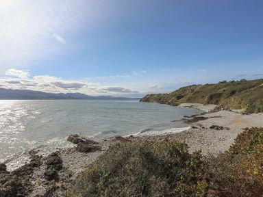 A rocky coastline with bushes and hills beside the sea under a partly cloudy sky at Flagstaff in Penmon near Beaumaris