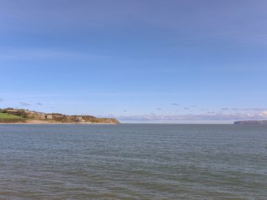 A view of the sea with a coastline featuring grassy cliffs and buildings under a clear sky at Flagstaff in Penmon near Beaumaris