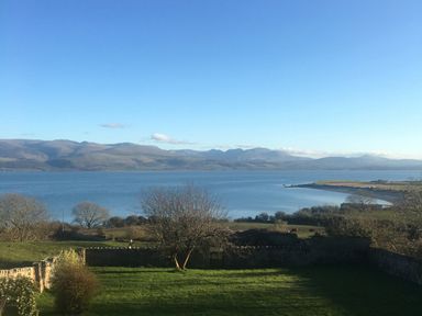 A view of a body of water with hills and mountains in the background and grassy land with trees in the foreground at Flagstaff in Penmon near Beaumaris