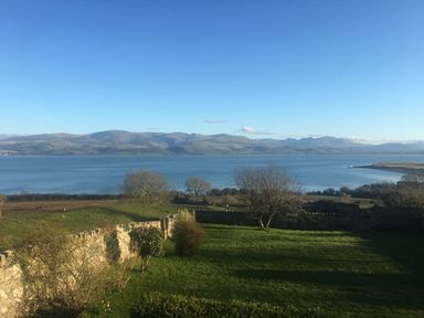 A waterfront view with grassy land and trees overlooking water and mountains at Flagstaff in Penmon near Beaumaris