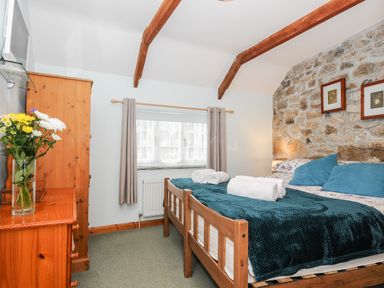 A bedroom with a wooden bed blue pillows and rolled towels on a blanket a wooden dresser with a vase of flowers a window with curtains and exposed wooden beams Mum's Cottage Minions near Upton Cross