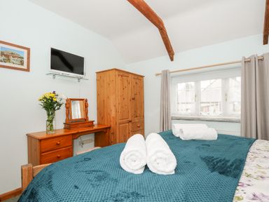 A bedroom with a wooden wardrobe desk with flowers and mirror a mounted TV and towels on a bed at Mum's Cottage in Minions near Upton Cross
