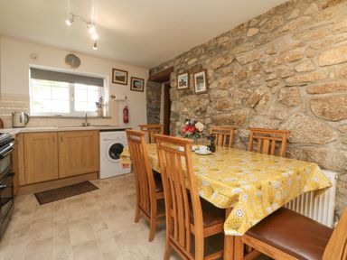 A kitchen with a wooden dining table covered with a yellow tablecloth and chairs with brown seats at Mum's Cottage in Minions near Upton Cross