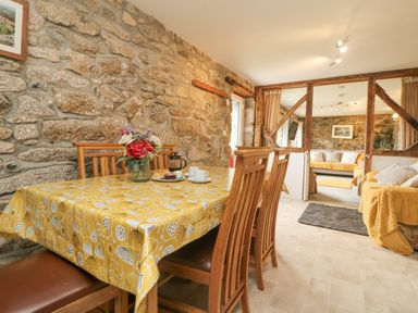 A dining area with wooden table and chairs with a yellow tablecloth and flowers on the table next to a sitting room with sofas and wooden beams at Mum's Cottage in Minions near Upton Cross