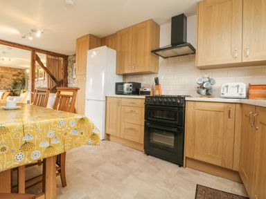 A kitchen with wooden cabinets a black oven a white refrigerator a microwave a toaster and a dining table with a yellow tablecloth at Mum's Cottage in Minions near Upton Cross