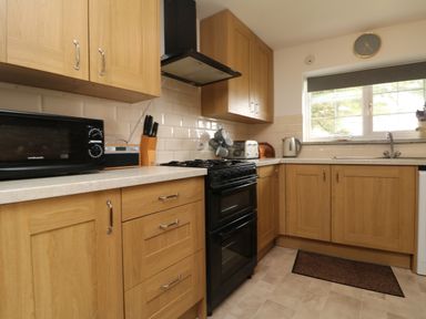 A kitchen with wooden cabinets a black oven a microwave toaster kettle and a window at Mum's Cottage Minions near Upton Cross