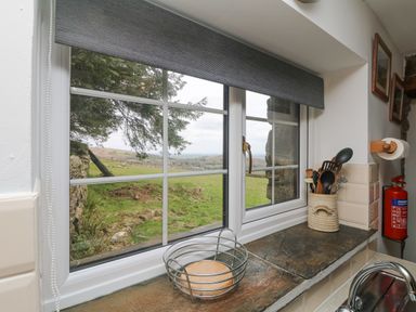 A kitchen window with a utensil holder and a fruit basket on the sill overlooking a grassy landscape at Mum's Cottage in Minions near Upton Cross