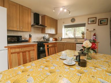 A kitchen with wooden cabinets a yellow patterned tablecloth and a table set with coffee cups and a flower vase at Mum's Cottage Minions near Upton Cross