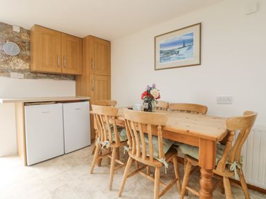 A dining area with wooden chairs and table a vase of flowers on the table wooden cabinets and two small refrigerators on the left wall a framed picture on the right wall at Bickle Bice Cottage in Minions near Liskeard