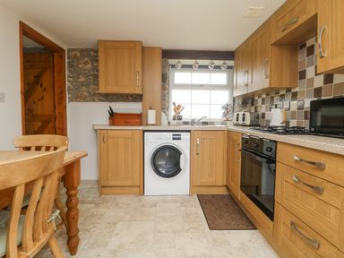 A kitchen with wooden cabinets a washing machine a stove toaster and a dining table with chairs at Bickle Bice Cottage in Minions near Liskeard
