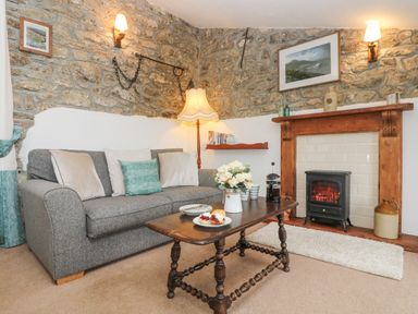 A living room with a gray sofa, wooden coffee table with flowers and plates, a fireplace with a stove and stone walls at Bickle Bice Cottage in Minions near Liskeard