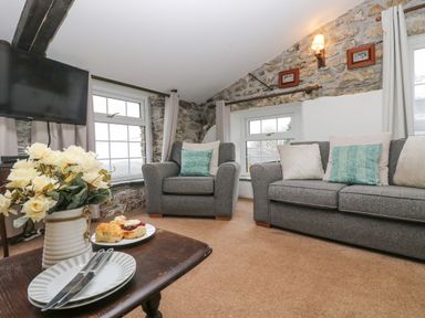 A living room with grey sofa and armchair with cushions a wooden table with plates and flowers exposed stone wall and windows at Bickle Bice Cottage in Minions near Liskeard