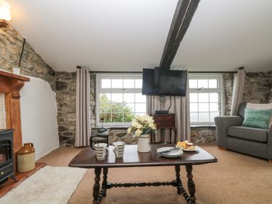 A living room with a wooden table holding cups and flowers a gray couch and a wall-mounted television at Bickle Bice Cottage in Minions near Liskeard