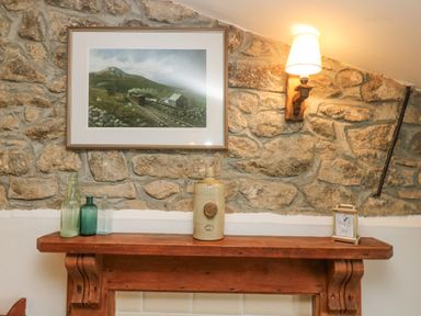 A stone wall with a wooden mantel holding glass bottles a ceramic jar and a clock below a framed picture of a train and a wall lamp at Bickle Bice Cottage in Minions near Liskeard