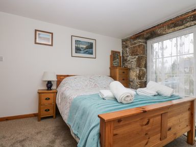 A bedroom with a wooden bed a bedside table with lamp a chest of drawers and a window with lace curtains at Bickle Bice Cottage in Minions near Liskeard