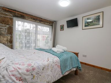 A bedroom with a bed with patterned bedding and towels on it a window with sheer curtains a wall-mounted tv and framed pictures at Bickle Bice Cottage in Minions near Liskeard