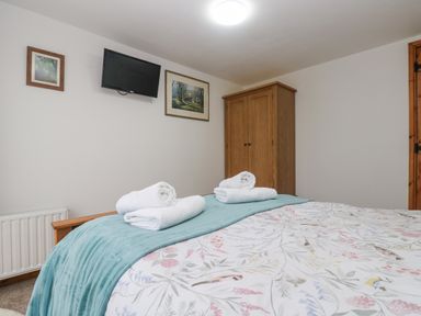 A bedroom with a bed covered with floral bedding and folded towels on a throw a wooden wardrobe a wall mounted TV and two framed pictures at Bickle Bice Cottage in Minions near Liskeard