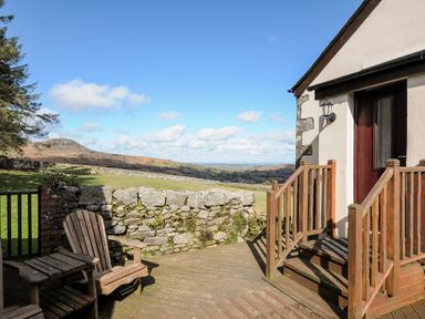 A wooden deck with chairs and stairs leading to a door overlooking a stone wall and grassy hills at Cheesewring Cottage in Minions near Upton Cross