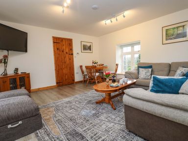 A living room with a gray couch a wooden coffee table a round wooden dining table and chairs a wooden door and a wall-mounted TV at Cheesewring Cottage in Minions near Upton Cross