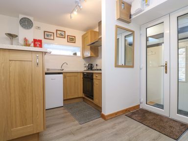 A kitchen area with wooden cabinets a small fridge, sink, oven, kettle, wall clock, and a glass door at Cheesewring Cottage in Minions near Upton Cross