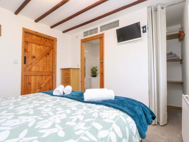 A bedroom with a bed covered by a patterned duvet and towels on top a wooden door a dresser a mounted television an open door to a bathroom and an open closet with shelves and hangers at Cheesewring Cottage in Minions near Upton Cross
