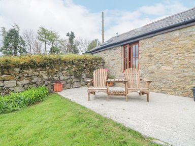 A patio with two wooden chairs and a table next to a stone wall and a house at Fizz-Gog Cottage Minions near Upton Cross