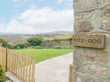 A stone wall with a sign saying Fizz-Gog next to a wooden gate and a grassy yard with hills in the background at Fizz-Gog Cottage in Minions near Upton Cross