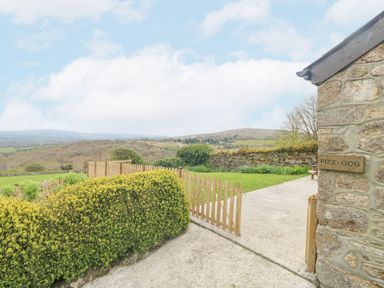 A stone wall with a sign saying Fizz-Gog a wooden gate opening to a garden with bushes and a concrete path with hills in the background at Fizz-Gog Cottage in Minions near Upton Cross