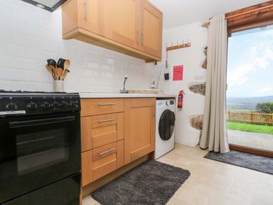 A kitchen with wooden cabinets a black oven a washing machine utensils in a container and a door with a view outside at Fizz-Gog Cottage Minions near Upton Cross