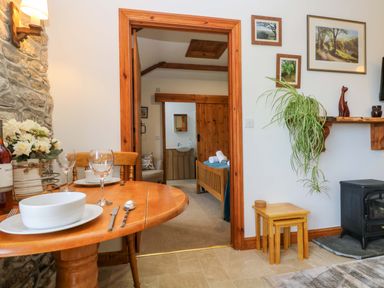 A dining area with a wooden table set with dishes and glasses next to a stone wall and an open doorway showing a bedroom with wooden furniture at Fizz-Gog Cottage in Minions near Upton Cross