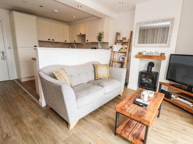 A living room with a gray sofa and wooden coffee table next to a fireplace and kitchen at 6 Cwrt Griffiths Prestatyn
