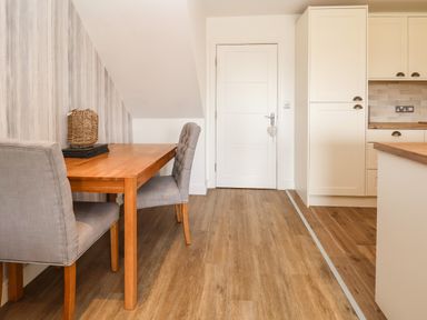 A dining area with a wooden table and two upholstered chairs next to a white door and kitchen cabinets at 6 Cwrt Griffiths in Prestatyn