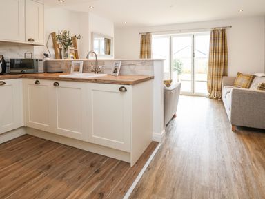 A kitchen with white cabinets and a wooden countertop opening to a living room with a gray sofa yellow plaid cushions and curtains at 6 Cwrt Griffiths in Prestatyn
