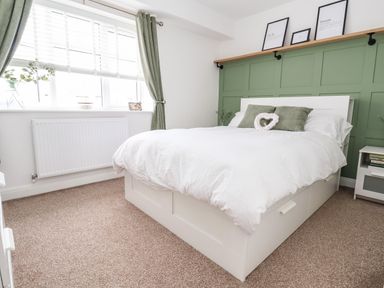 A bedroom with a white bed and green pillows next to a window with green curtains at 6 Cwrt Griffiths in Prestatyn