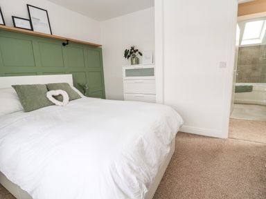 A bedroom with a white bed and green pillows and a green paneled wall with framed pictures on a shelf and a white chest of drawers next to an open door leading to a bathroom at 6 Cwrt Griffiths in Prestatyn