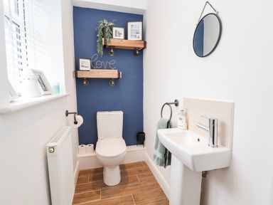 A bathroom with a toilet and a sink a mirror hanging above the sink wooden shelves with decorations on a blue accent wall at 6 Cwrt Griffiths in Prestatyn