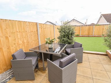 An outdoor patio area with a glass top table and four grey chairs next to a wooden fence and green lawn at 6 Cwrt Griffiths in Prestatyn