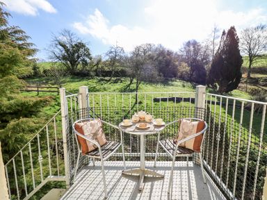 A small balcony with a metal table and two chairs with cushions overlooking a green grassy area with trees at The Cheese Loft in White Mill near Carmarthen