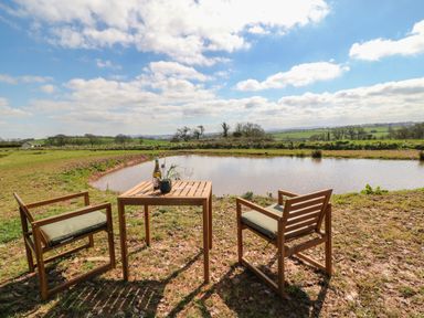 An outdoor seating area with a wooden table and two chairs next to a pond in a rural setting at Tawny in Morchard Bishop
