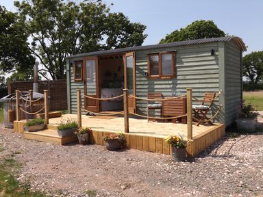A small wooden cabin with open double doors showing a bed inside a wooden deck with two chairs a table and flower pots at Tawny in Morchard Bishop