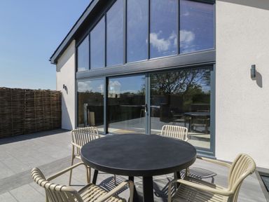 An outdoor patio with a round black table surrounded by beige chairs in front of a building with large windows at The Barn in Llanfechell