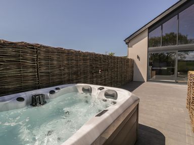 An outdoor patio with a hot tub and a wicker fence next to a modern building with large windows at The Barn in Llanfechell