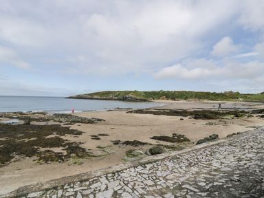 A sandy beach with rocks and a stone pathway near a calm sea with a green hill in the background at The Barn in Llanfechell