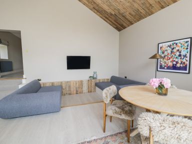 A living room with two gray sofas a round wooden table with flowers chairs with fur covers and a wall-mounted television at The Barn in Llanfechell
