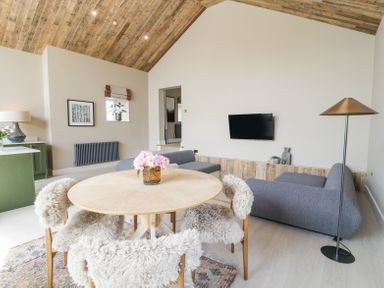 A living room with a round wooden table and chairs with fur covers two gray sofas a wall mounted TV and a floor lamp at The Barn in Llanfechell