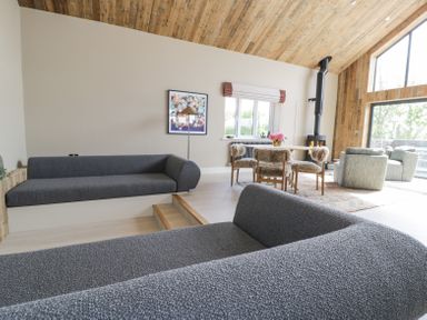 A living room with grey sofas a dining table with chairs a wood stove and large windows at The Barn in Llanfechell