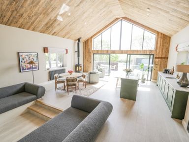 A living room and kitchen with wood-paneled ceiling large windows and grey seating at The Barn in Llanfechell