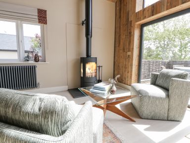 A living room with two patterned armchairs a glass coffee table with books a wood stove and large window with outdoor view at The Barn in Llanfechell