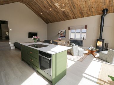 An open plan living room and kitchen with a green island and stove a dining table with chairs two armchairs a wood stove and a wooden ceiling at The Barn in Llanfechell