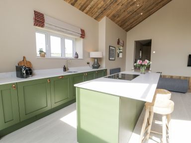 A kitchen with green cabinets, a white countertop island with stools and a pink flower vase at The Barn in Llanfechell